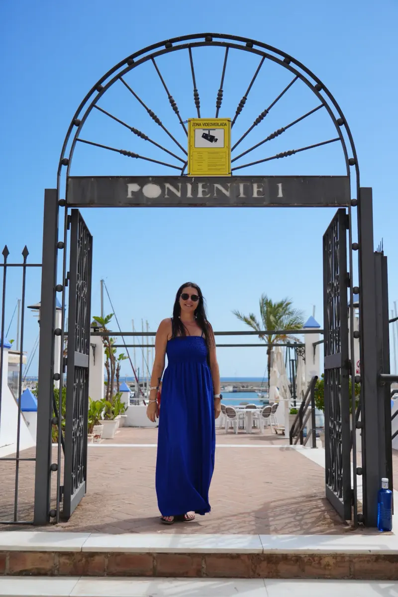 Heidi standing under the Poniente 1 iron gate at Estepona port, where the Sunday marina market sets up