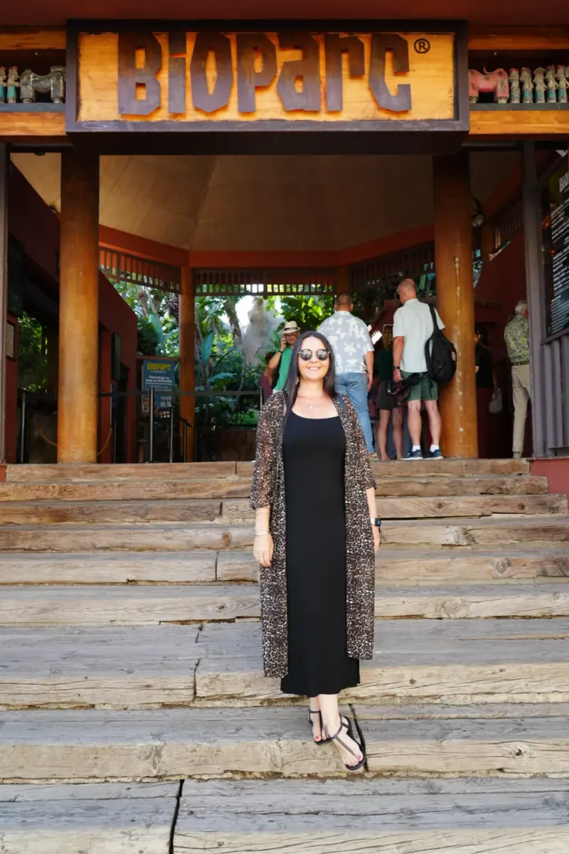 Heidi in sunglasses, a leopard-print cardigan and a long black dress, standing on the wooden steps under the Bioparc Fuengirola entrance sign