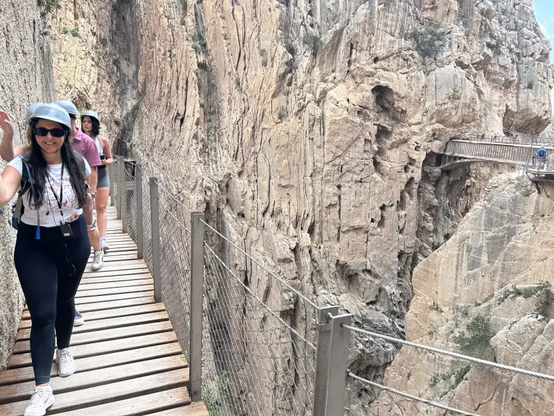 Heidi in a grey helmet and sunglasses smiling and waving on a Caminito del Rey boardwalk, with friends behind her and the gorge walls beyond