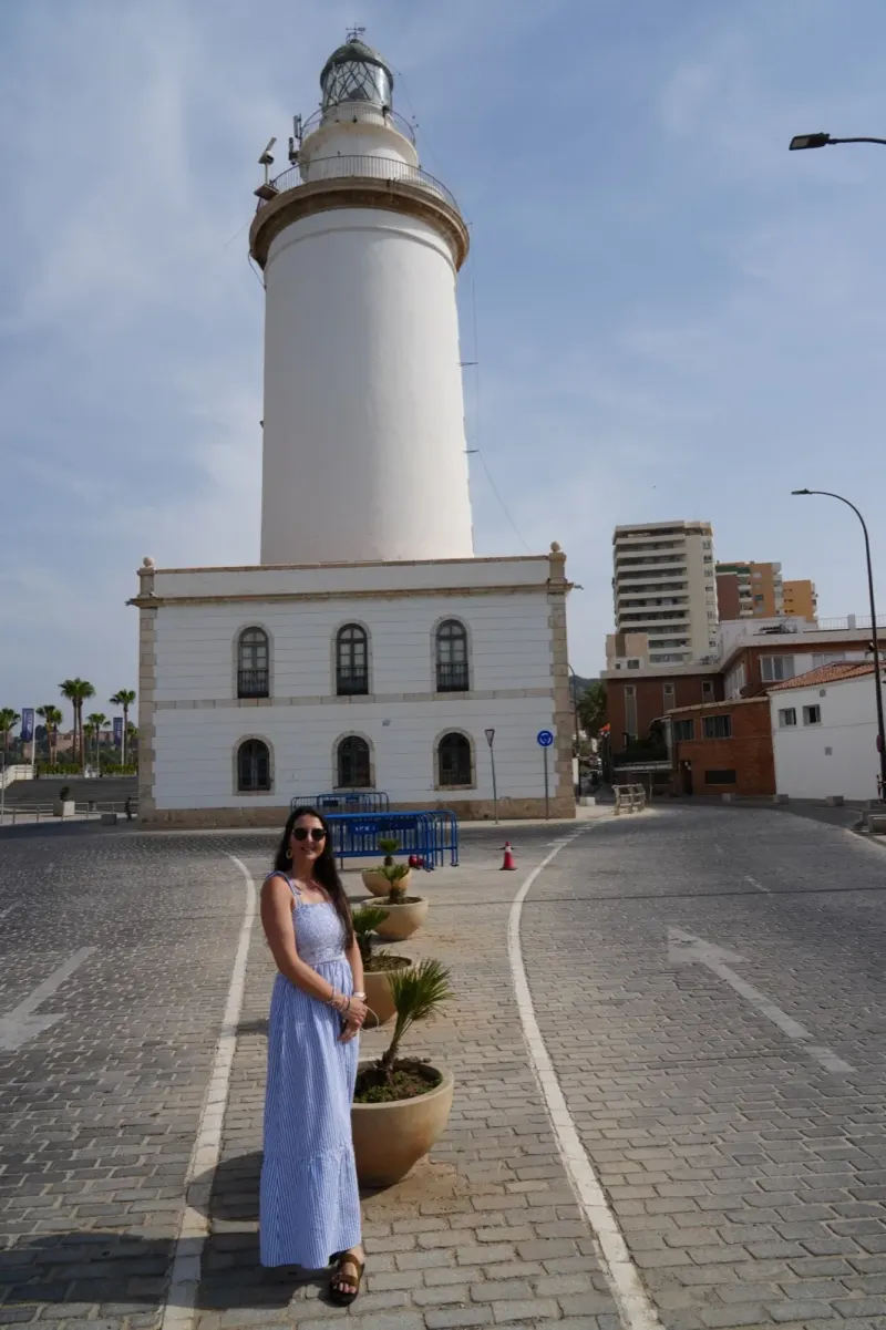 Heidi in a long blue-and-white dress standing by the white La Farola lighthouse in Málaga