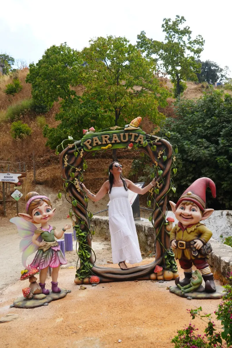 Heidi in a white sundress standing inside the carved wooden Parauta arch, with a fairy statue on the left and a red-hatted gnome statue on the right