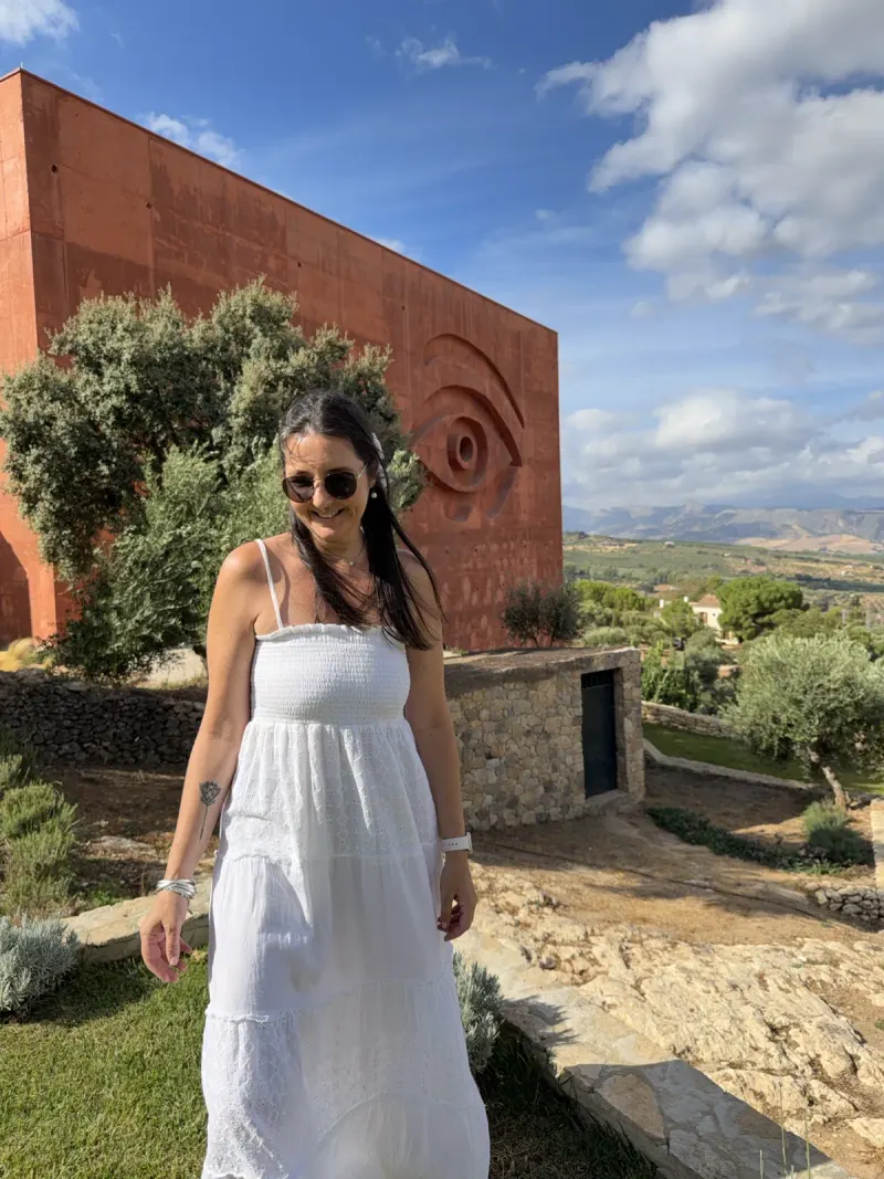 Heidi in a white sundress smiling in front of the red cube of LA Almazara near Ronda, with the huge carved eye on its wall and olive groves and mountains behind
