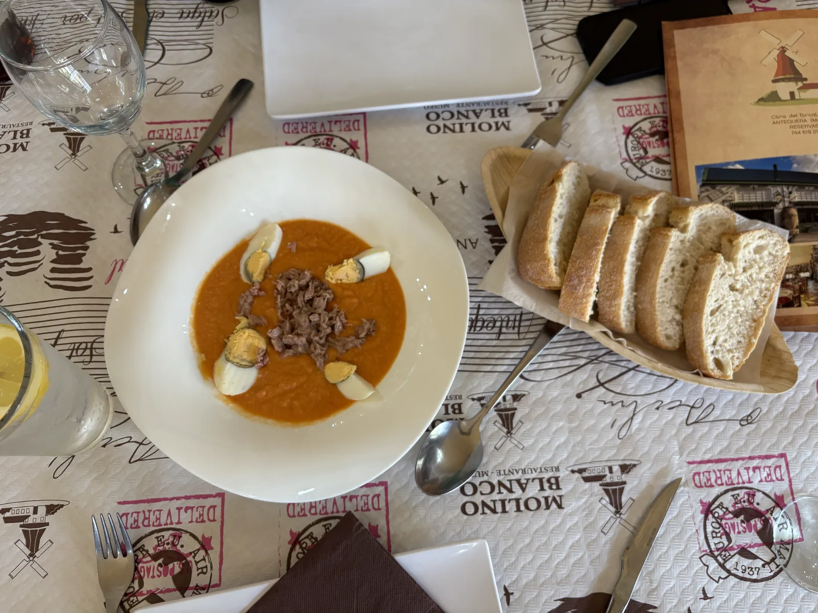 A bowl of porra antequerana topped with tuna and hard-boiled egg, served with a basket of bread at a restaurant table in Antequera
