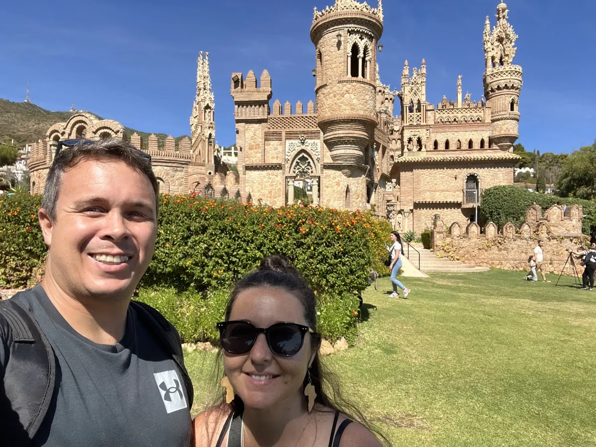 Heidi and Jethro taking a selfie on the lawn in front of Castillo de Colomares, its stone turrets and towers behind them and orange flowers on the hedges