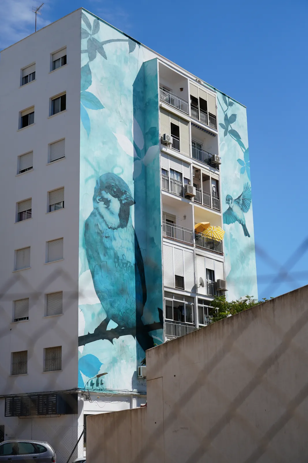 Giant teal sparrow mural wrapping around an apartment building corner in Estepona