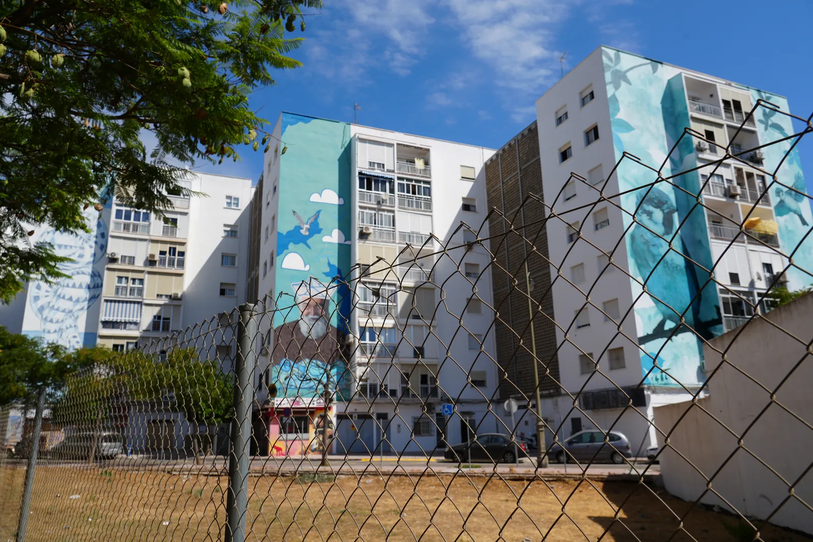 Wide street-level view of an apartment block mural glimpsed above the trees in a pedestrian square in Estepona