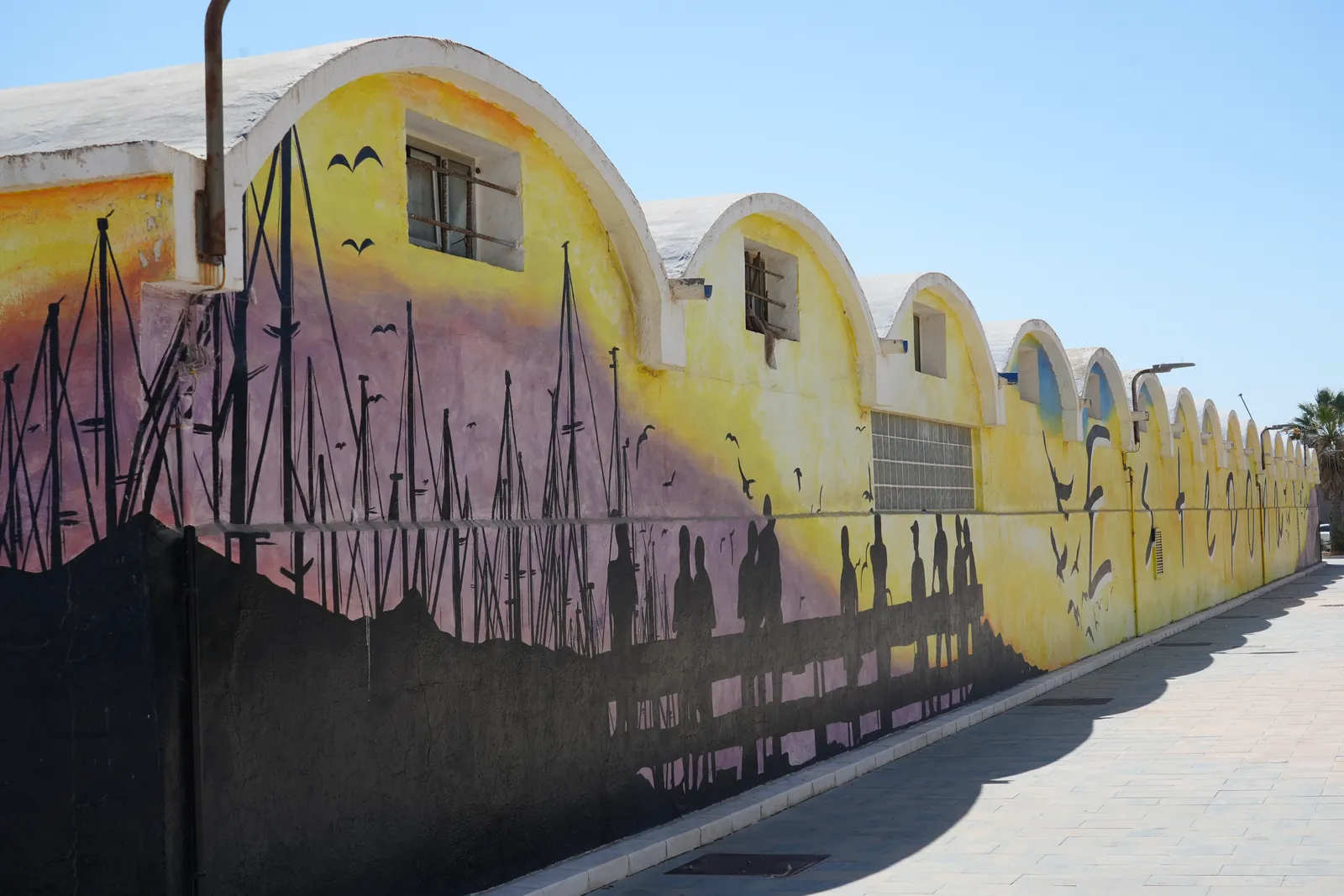 A curved harbourside wall in Estepona's fishing port painted with fishermen silhouettes against a sunset sky