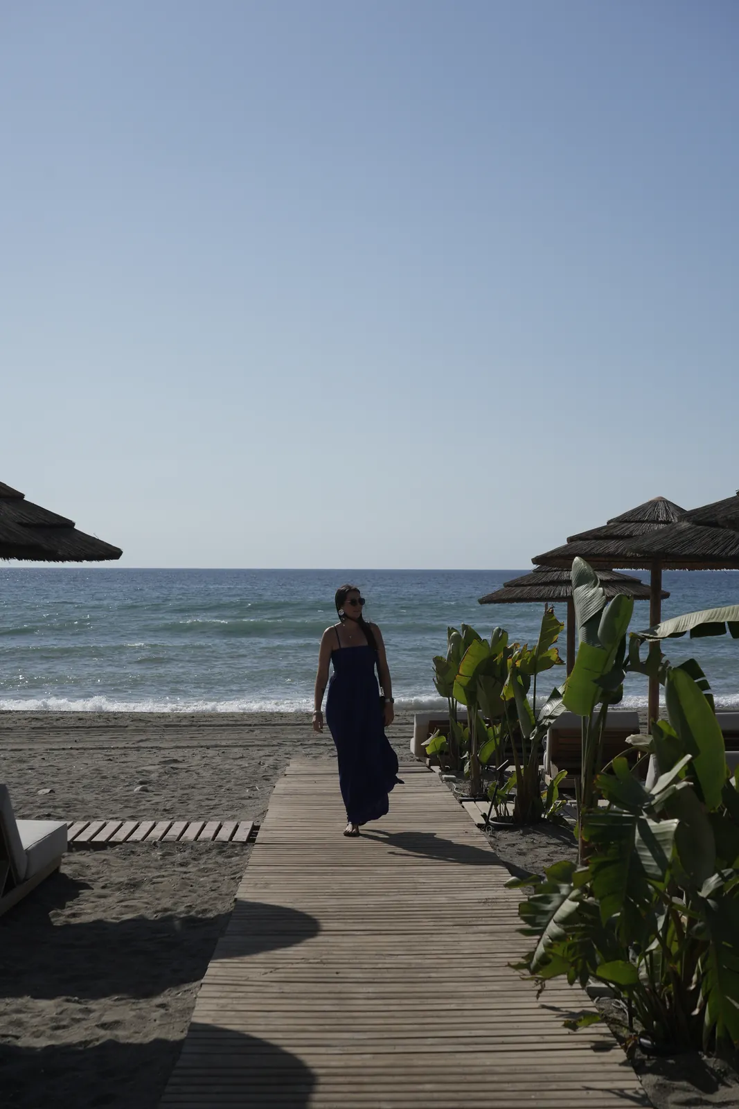 Heidi walking along a wooden boardwalk between straw umbrellas and banana leaf plants at the beach in Estepona