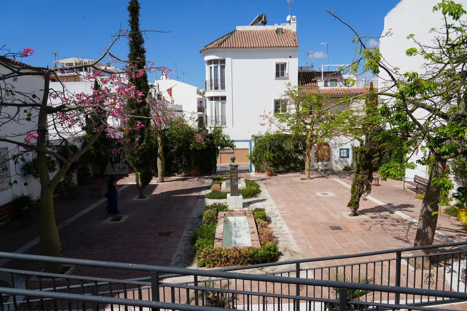 Wide view of Plaza de Antonio Gala in Estepona, the bronze bust on its pedestal, a rectangular fountain, benches and silk floss trees in bloom