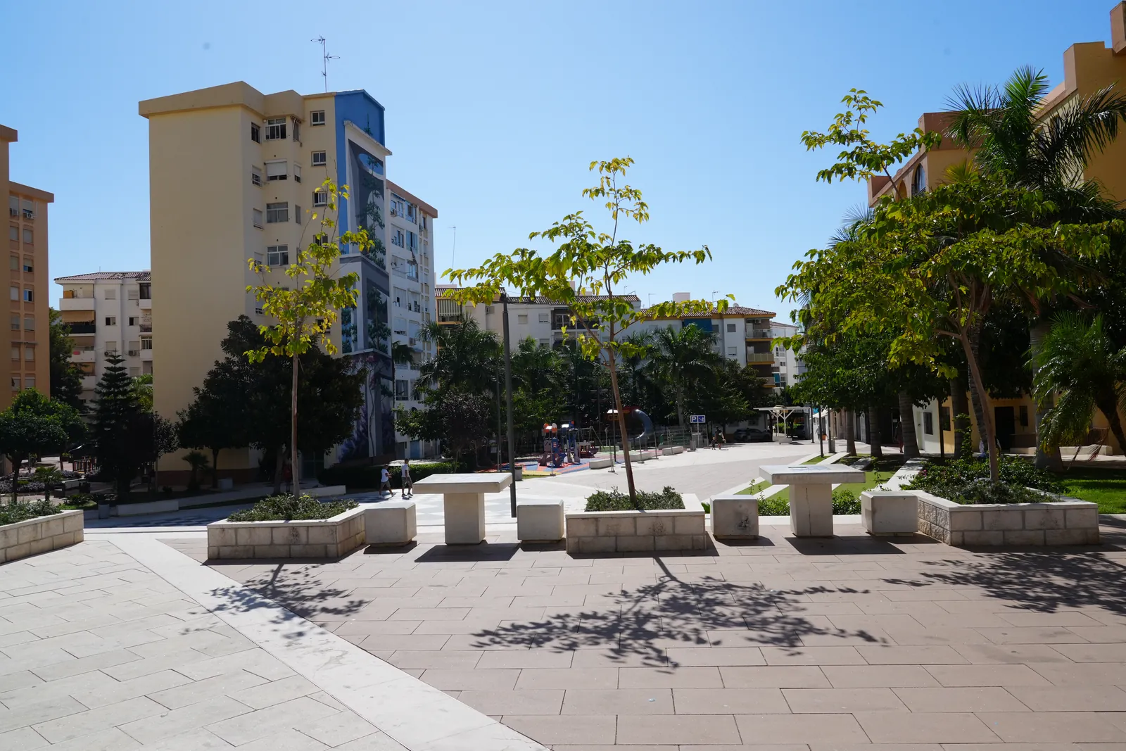 Wide view of a pedestrian square in Barriada Primavera, Estepona, palm trees and benches with an apartment block mural visible in the background