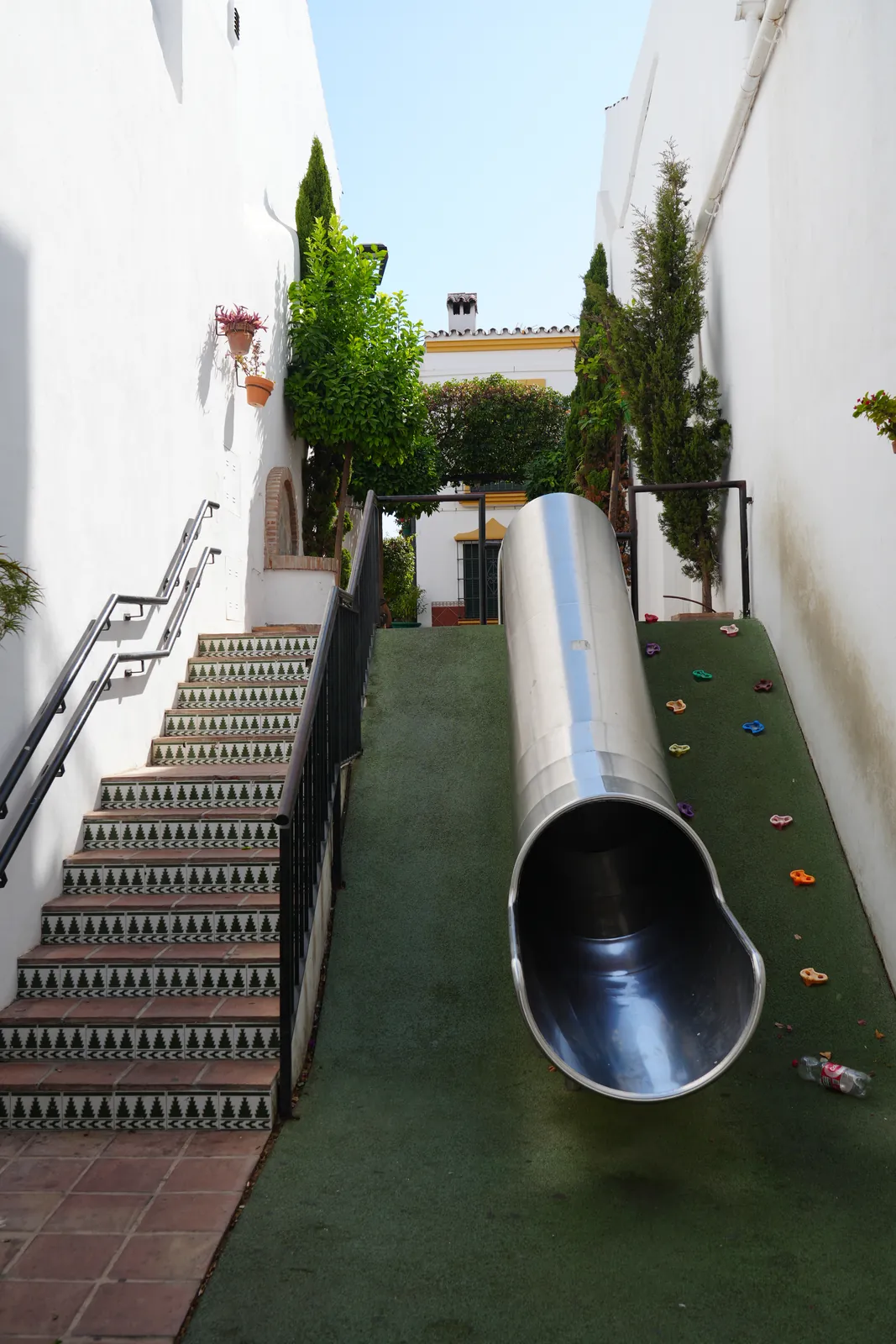 Stainless-steel tube slide built alongside a tiled staircase in Pasaje Manuel Sánchez Bracho, Estepona old town, with coloured climbing holds set into the turf incline beside it