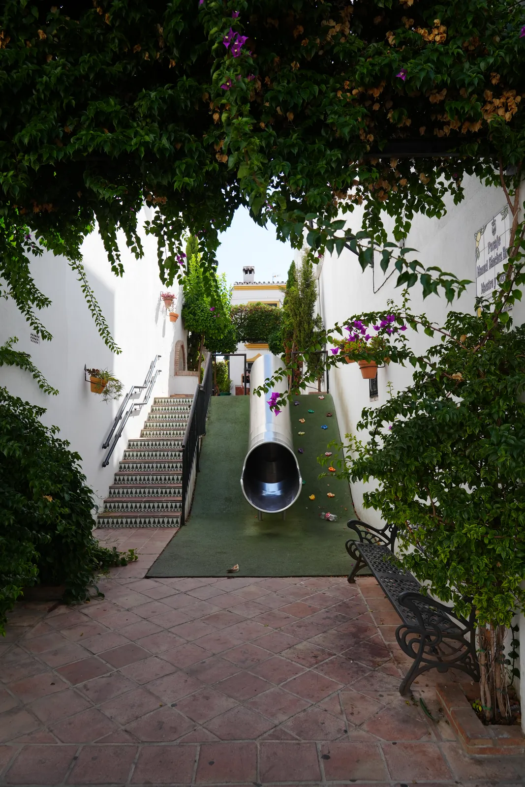 A stainless-steel tube slide bolted into the staircase at Pasaje Manuel Sánchez Bracho, bougainvillea overhead, Estepona old town