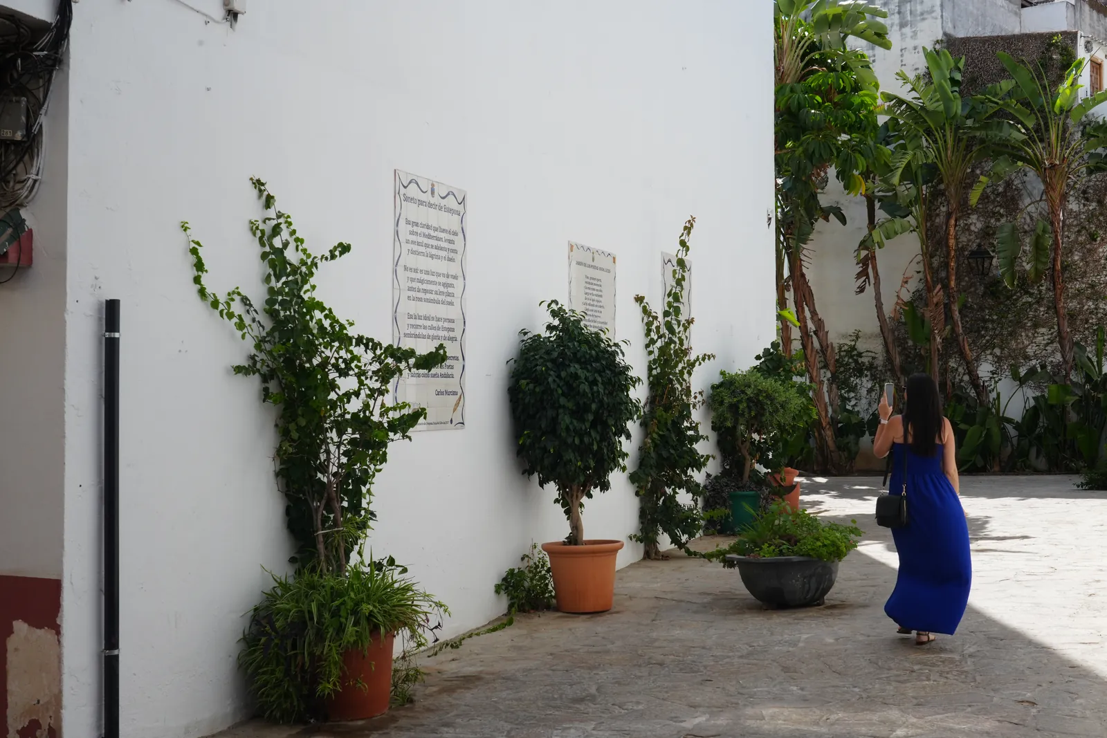 Heidi photographing the ceramic poem tiles in the Jardín de los Poetas Andaluces passage in Estepona, surrounded by potted plants and palms