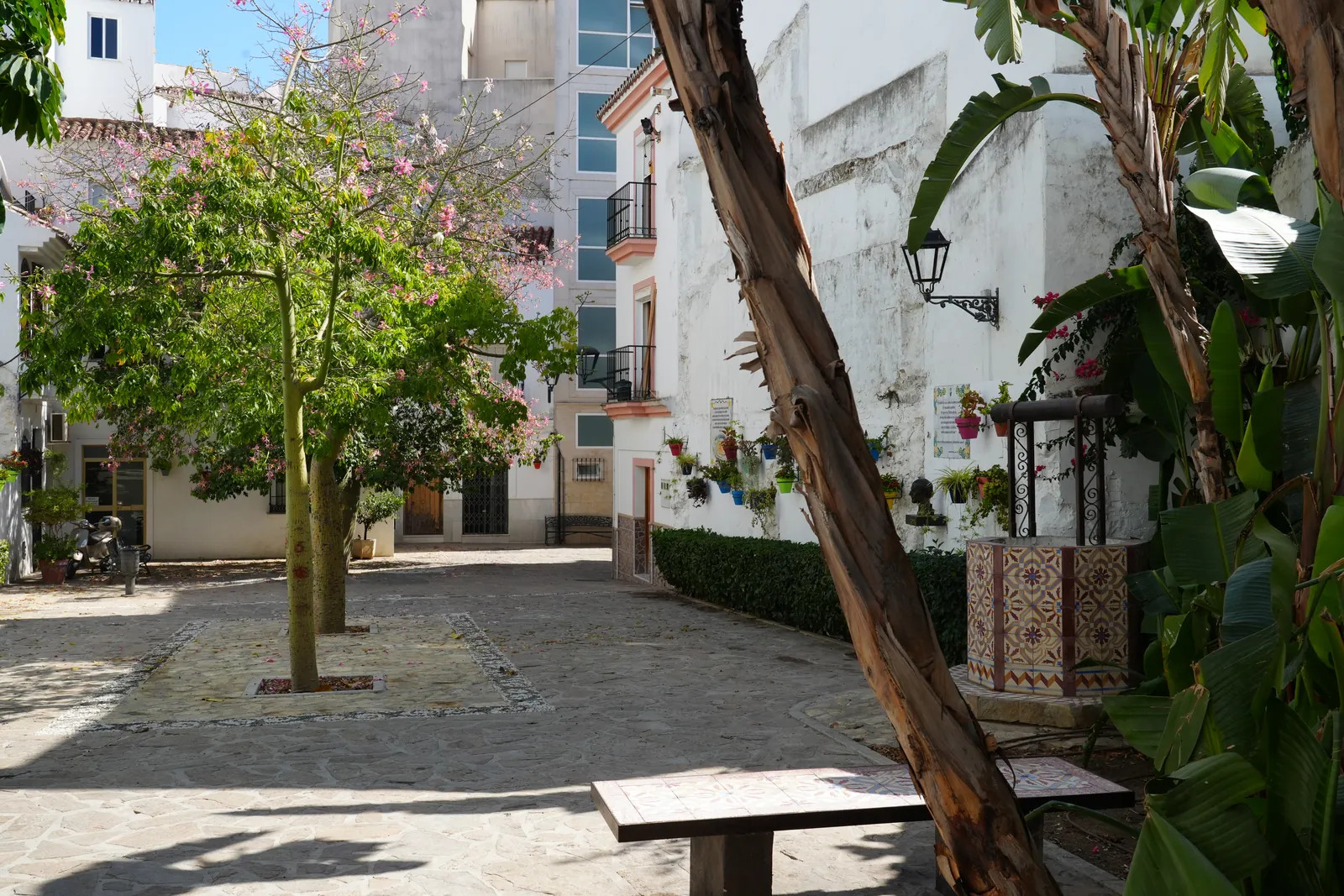 Wide view of the courtyard in the Jardín de los Poetas Andaluces in Estepona, a flowering tree, a tiled well and hanging flowerpots along the whitewashed wall