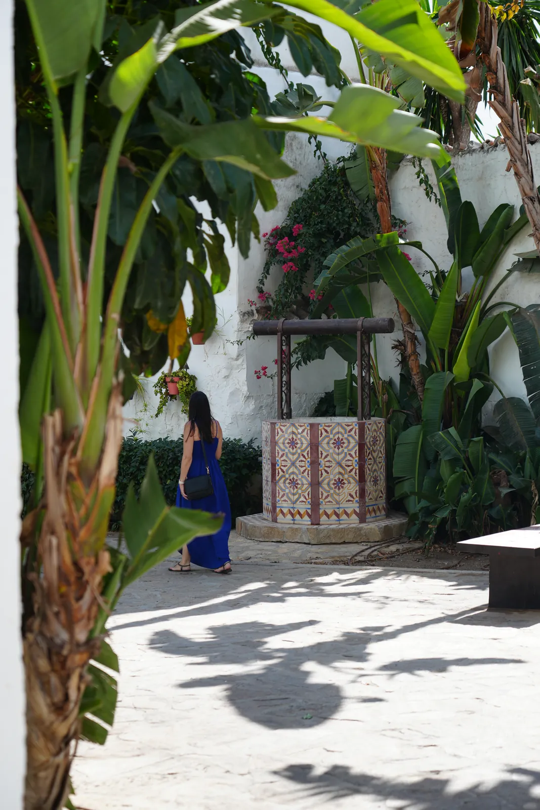 Heidi walking away through the Jardín de los Poetas Andaluces towards the tiled well, framed by banana leaves