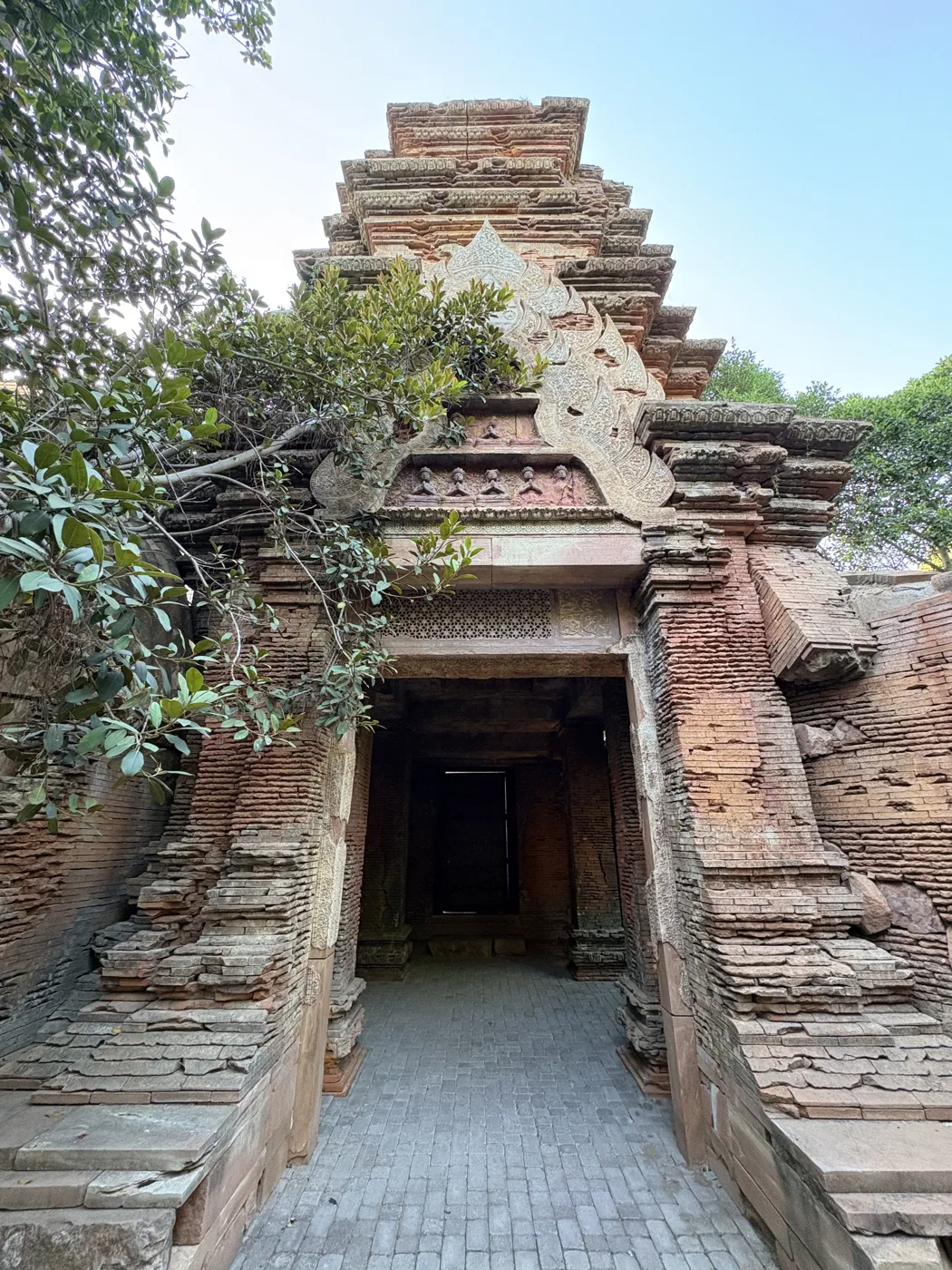 A stepped red-brick Khmer-style temple gateway with a carved pediment and trees growing over it at Bioparc Fuengirola