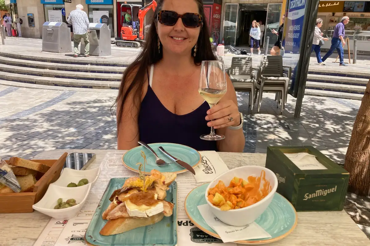 A woman holding a glass of cava with a table full of tapas, including patatas bravas and olives.
