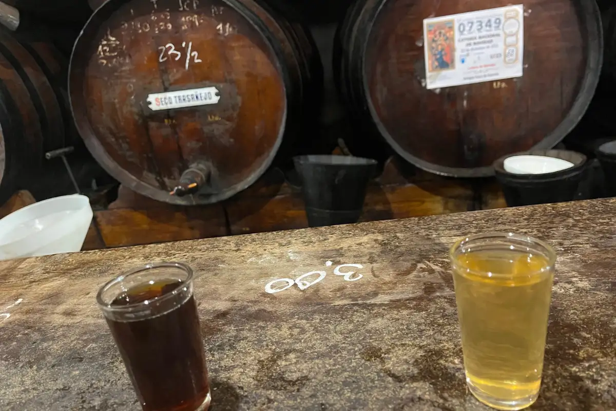 Two small glasses of Málaga wine on the worn wooden counter at Antigua Casa de Guardia, the tab chalked on the wood between them and the barrels behind