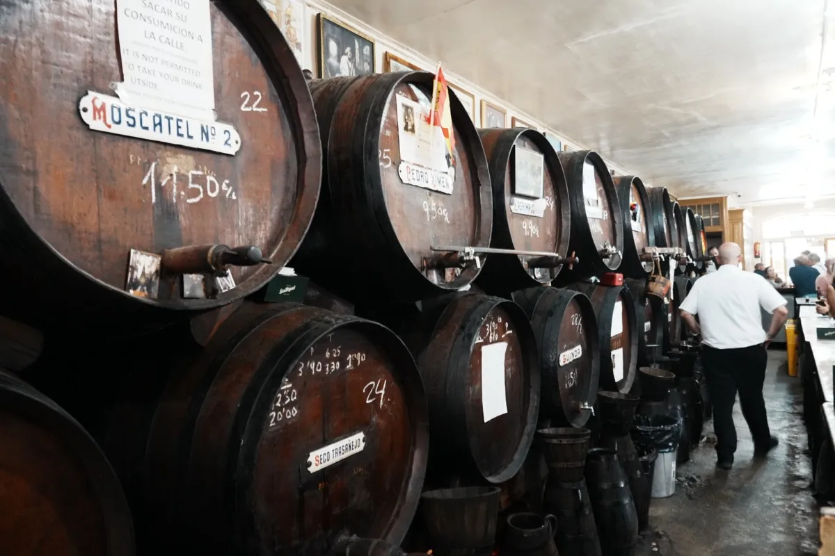 Dark wine barrels stacked behind the bar at Antigua Casa de Guardia, a Moscatel label and prices chalked on the wood, a barman in a white shirt beyond