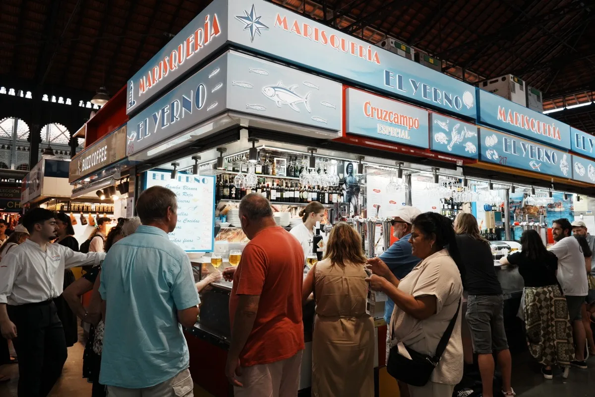 People packed around the seafood counter of Marisquería El Yerno inside Atarazanas market, its blue signs above the bar and glasses of beer under the market's iron roof