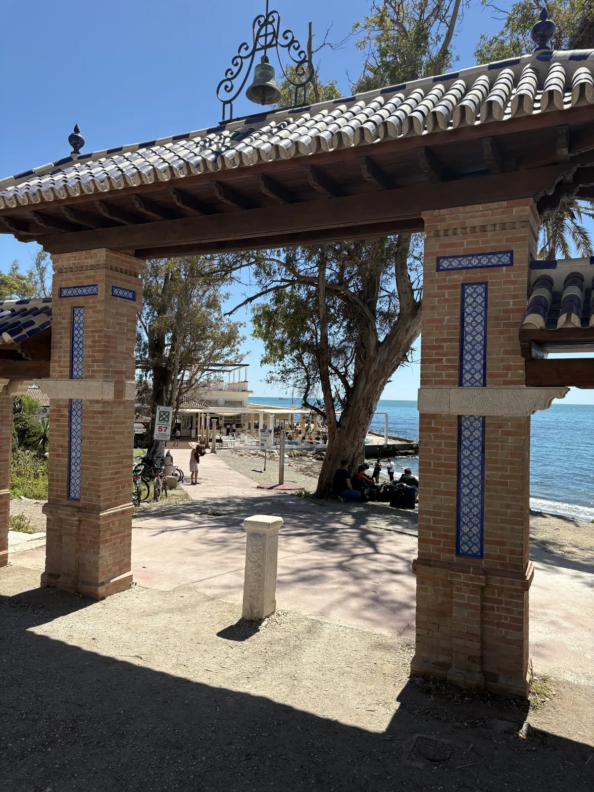 The brick and tile entrance gate to Baños del Carmen, a bell on top, with the eucalyptus, the sea and El Balneario's white pavilion through the arch