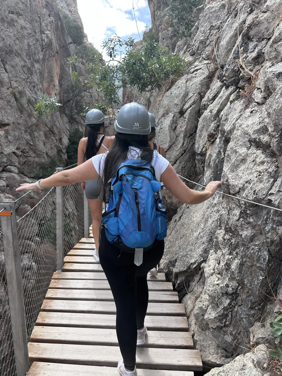 Heidi from behind in a helmet, one hand on the rail and one on the rock, on a narrow boardwalk on the Caminito del Rey