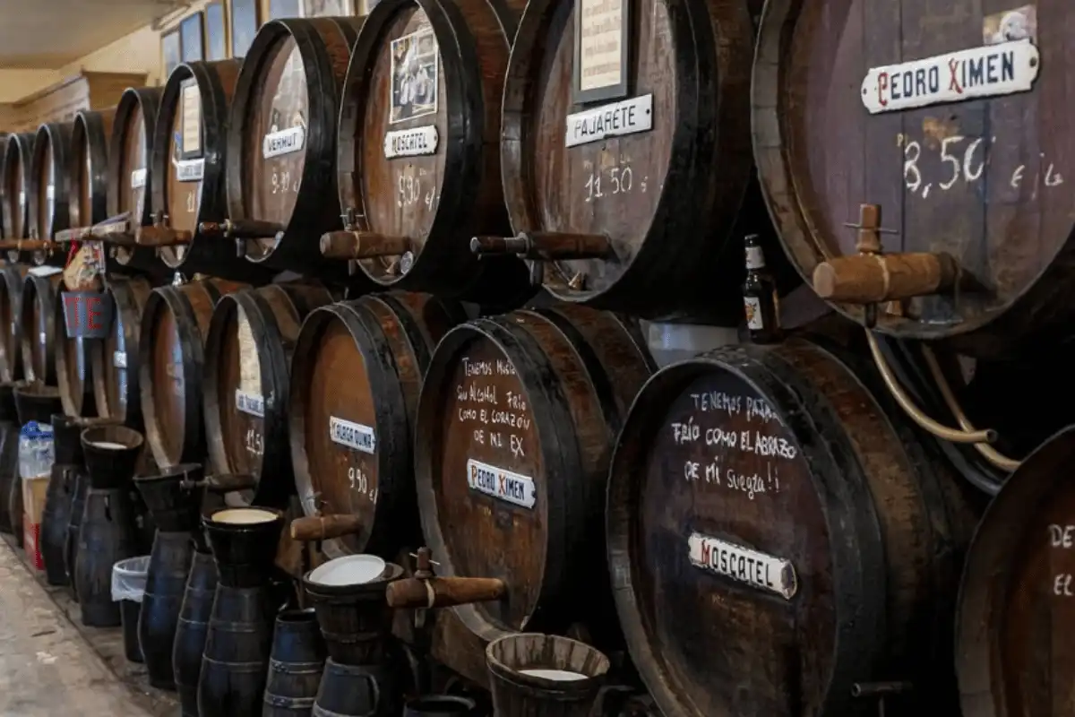 Wall of dark wooden barrels labelled Pedro Ximén, Pajarete and Moscatel, with prices chalked on the wood, behind the counter at Antigua Casa de Guardia, Málaga's oldest bar