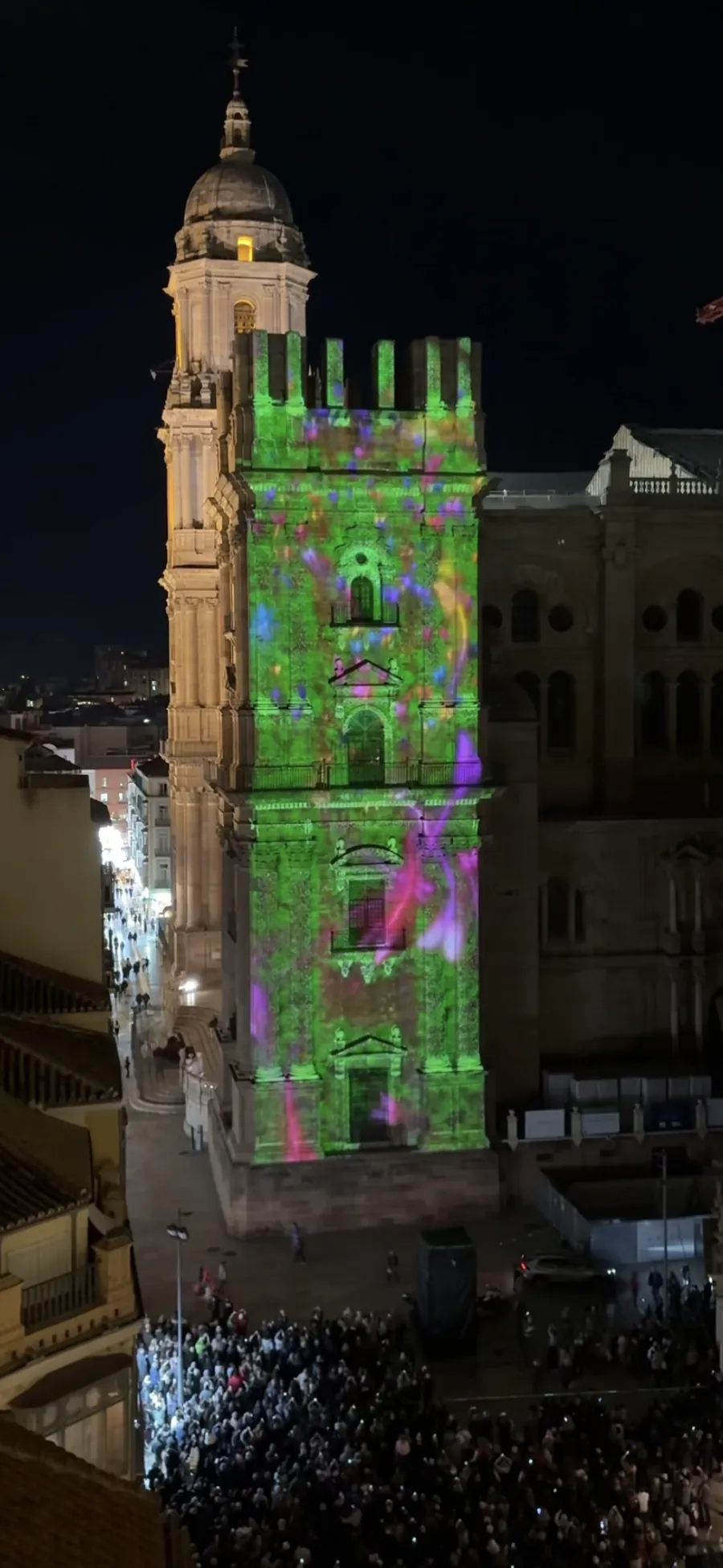 Green and purple video mapping light show projected onto Malaga Cathedral's tower, seen from above a packed crowd