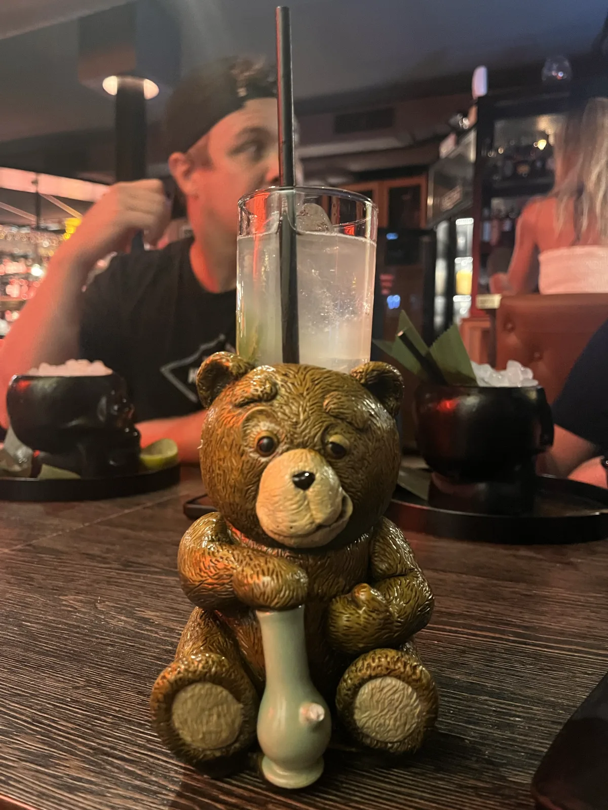 A cocktail at Chester & Punk served in a ceramic teddy bear clutching a tiny bong, the tall glass and black straw rising out of its head, skull-shaped cups on the table behind