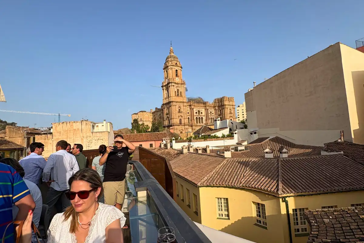 A crowd at the railing of the Chinitas rooftop bar with glasses in hand, Málaga Cathedral's tower behind them in the evening light