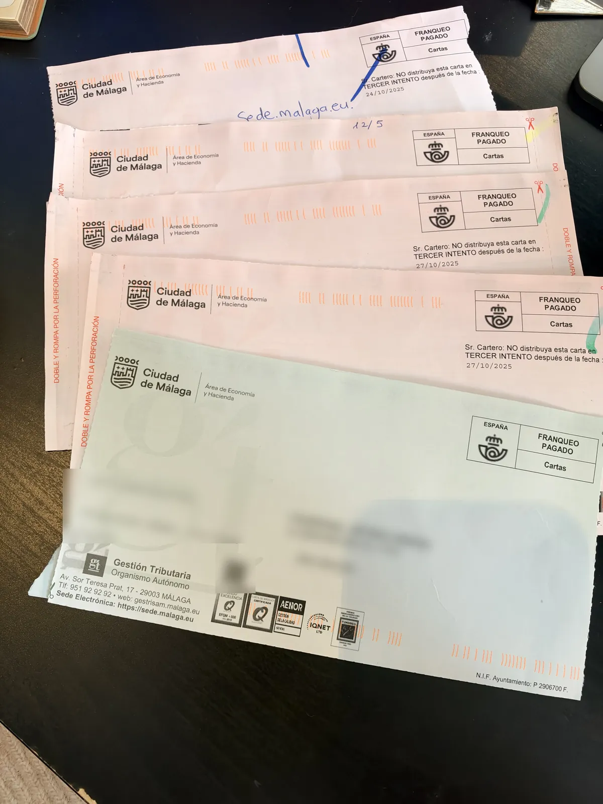 Four pink fine envelopes from the Ciudad de Málaga and one green envelope from its tax office, Gestión Tributaria, fanned out on a dark table, with the name and address blurred