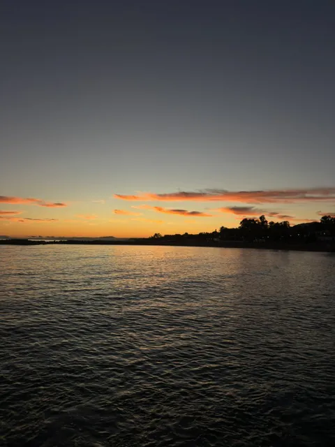 Calm sea at dusk along Málaga's eastern beaches, with a breakwater and a tree-lined point silhouetted against the last orange clouds