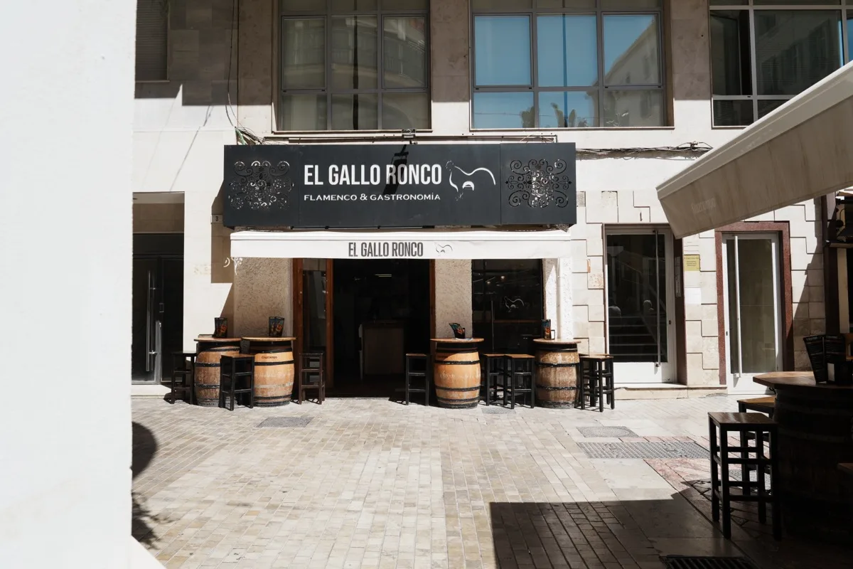 The black El Gallo Ronco flamenco and gastronomía sign above the entrance on Plaza de las Flores, wine barrels standing outside the door