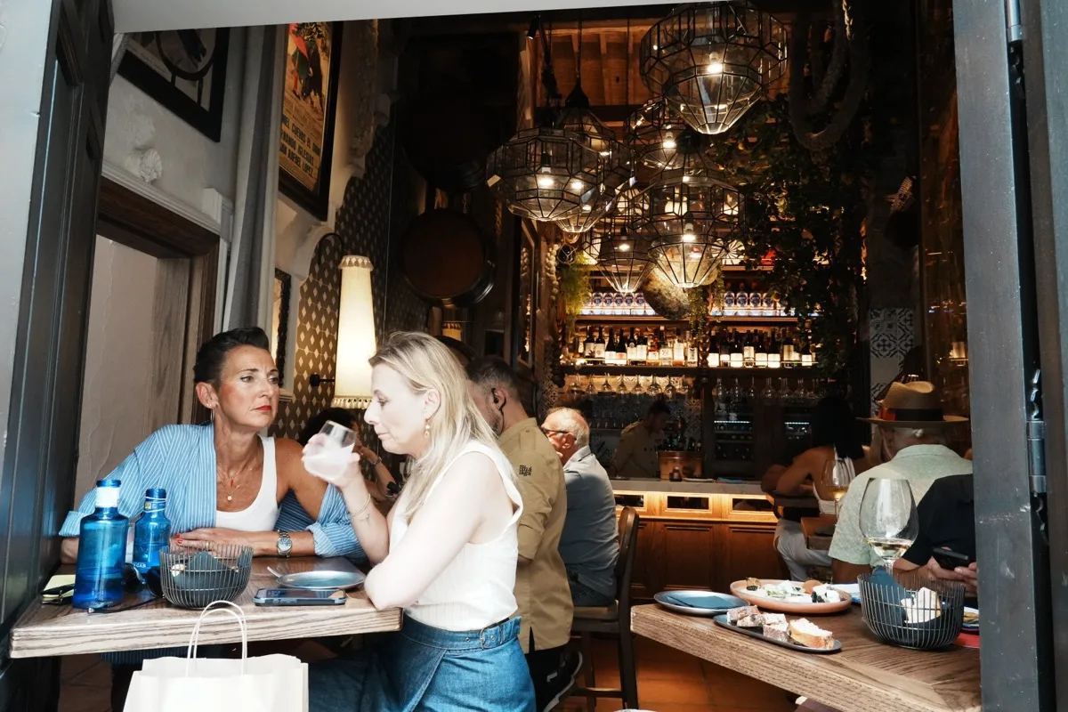 Two women sharing a drink at a table inside El Pimpi, geometric glass lamps and hanging plants over the bar behind them