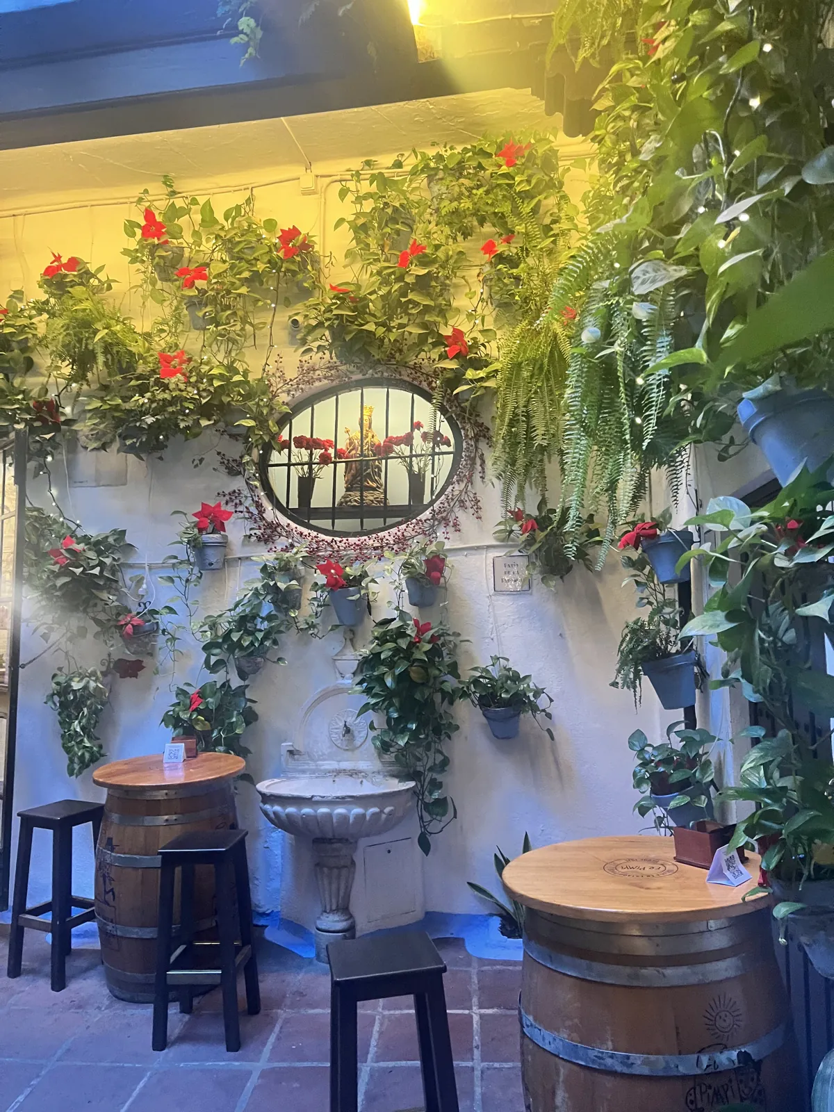 El Pimpi's patio: a white wall hung with plants and red poinsettias around a barred niche with a statue of the Virgin, a stone basin below and barrel tables with black stools