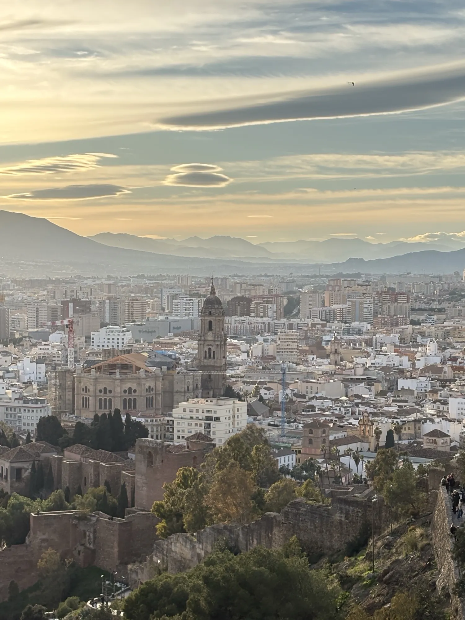 View west over Málaga from the Alcazaba walls, with the cathedral tower and the mountains beyond lit in warm dusk light