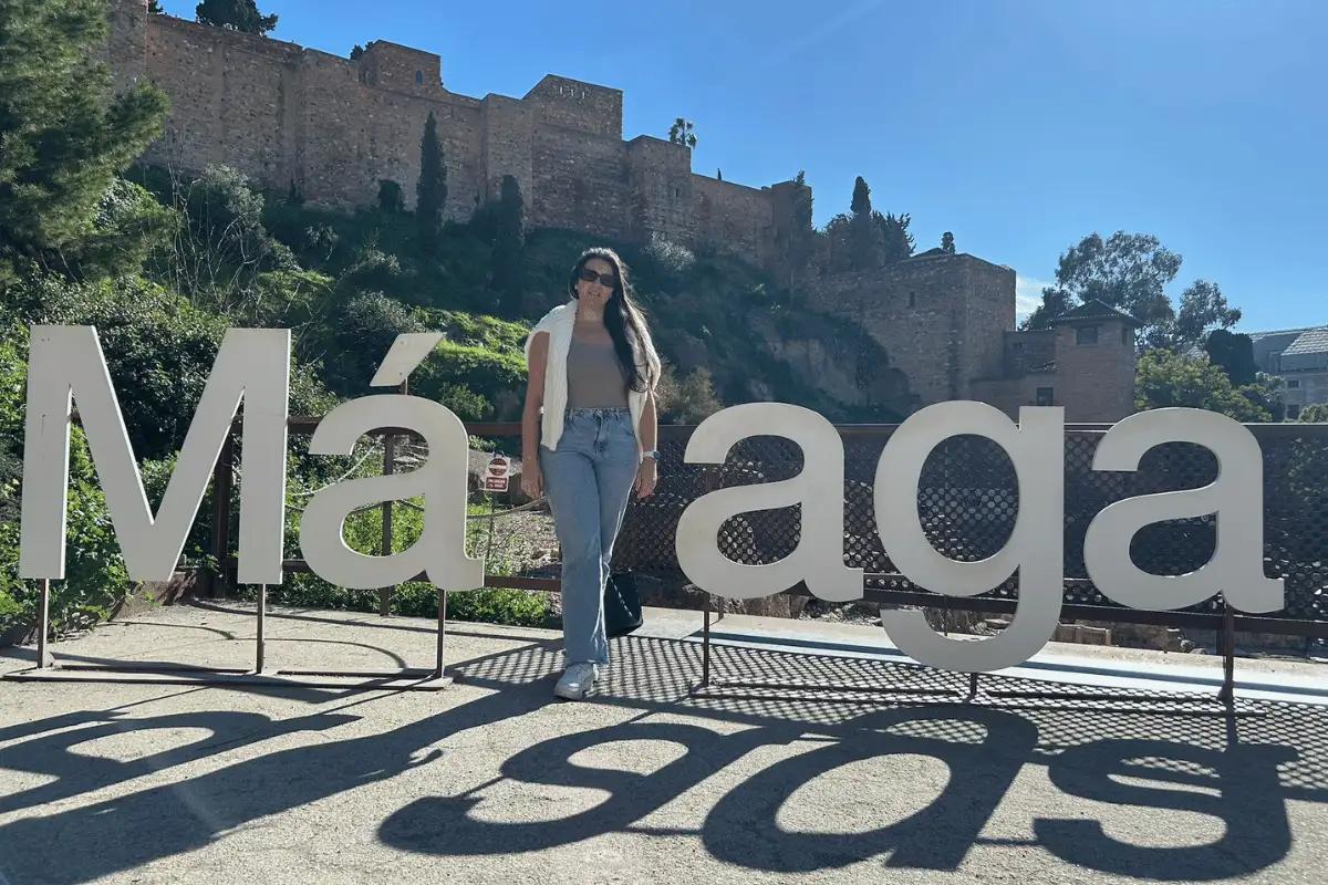 Heidi posing at the Malaga sign with the Roman Theatre and Alcazaba behind her