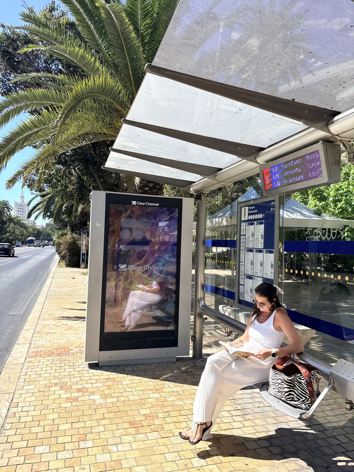 Heidi reading on the bench at the Parque Rectorado bus stop in Málaga, with the arrivals board showing Lines 3, 11 and 32