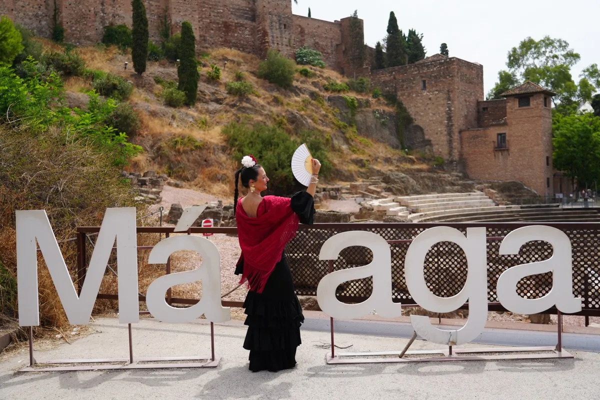 Heidi in a black flamenco dress and red fringed shawl, fan raised, standing in front of the white Málaga letters with the Roman Theatre and Alcazaba behind her during the Feria de Málaga