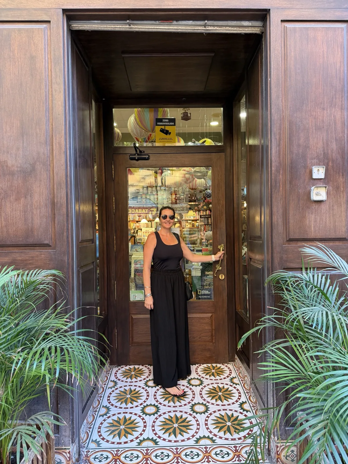 Heidi smiling in the doorway of Mapas y Compania in Malaga, hot air balloon decorations visible inside, standing on a patterned tiled entrance