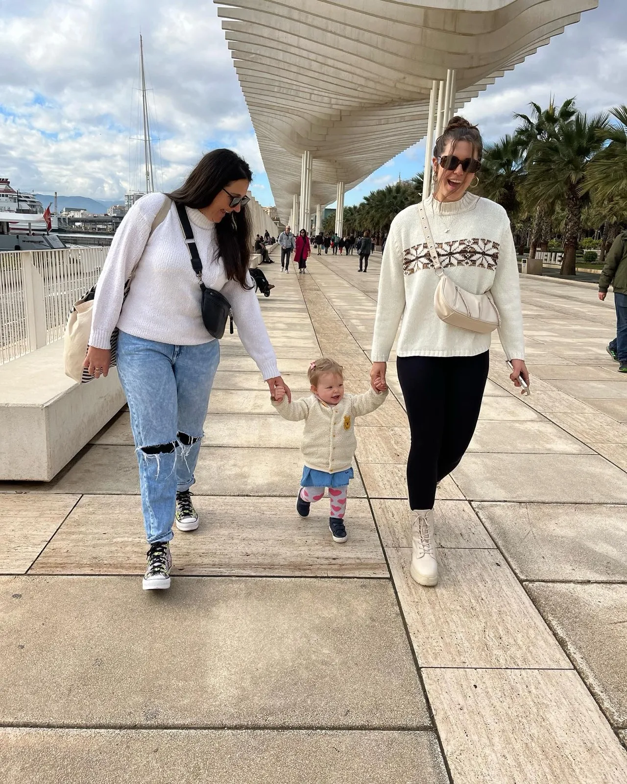 Heidi walking hand in hand with a toddler along the Muelle Uno promenade under the wooden pergola in Málaga on a cool November day