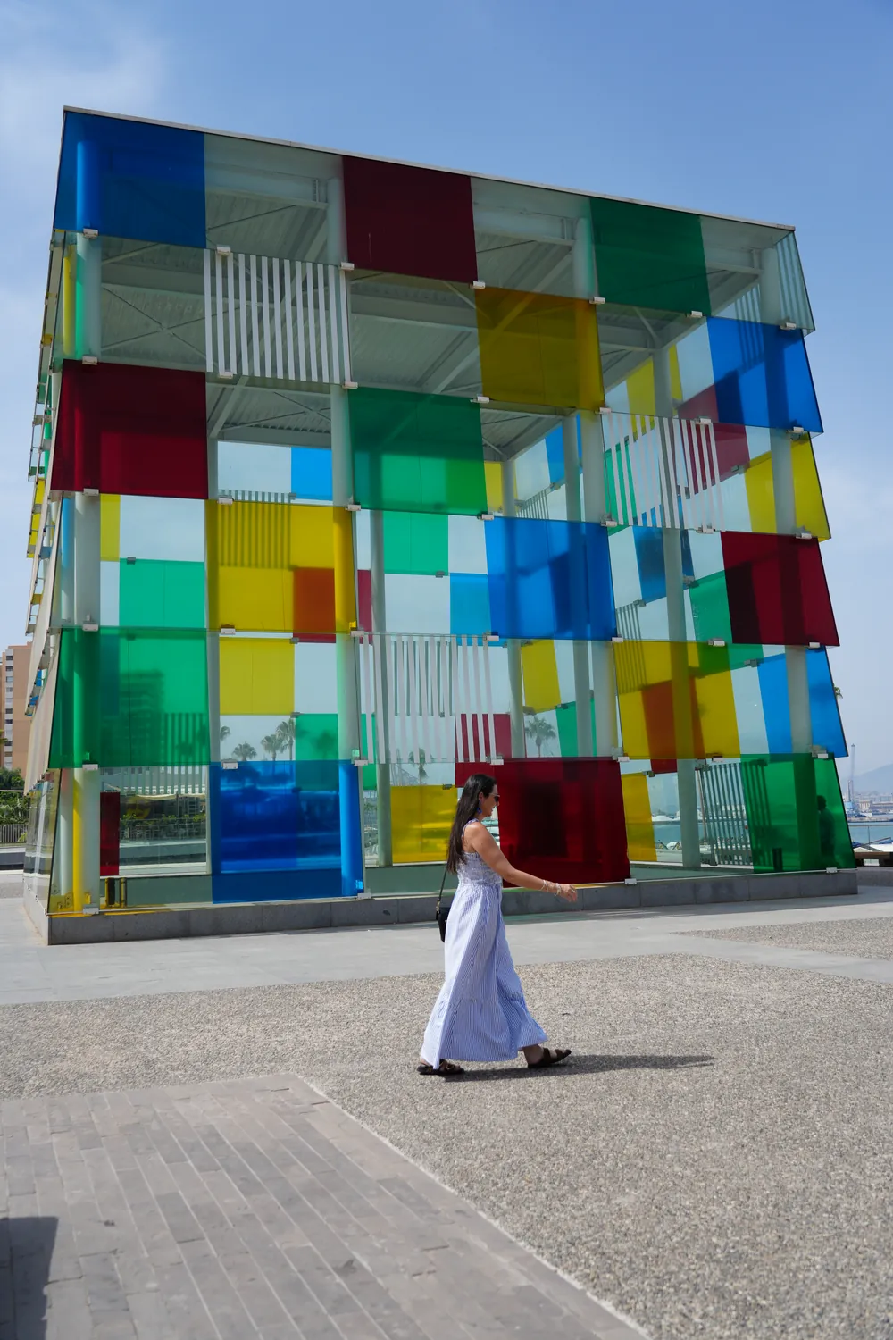 Heidi in a blue striped maxi dress walking past the multicoloured glass cube of the Centre Pompidou on Muelle Uno in Málaga