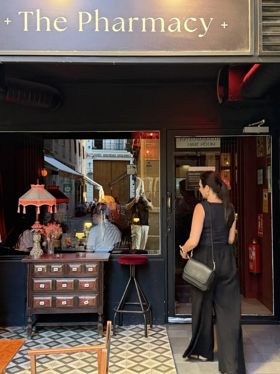 Heidi photographing the reflection in the window of The Pharmacy speakeasy in Málaga at dusk, the lit sign and apothecary cabinet visible through the glass