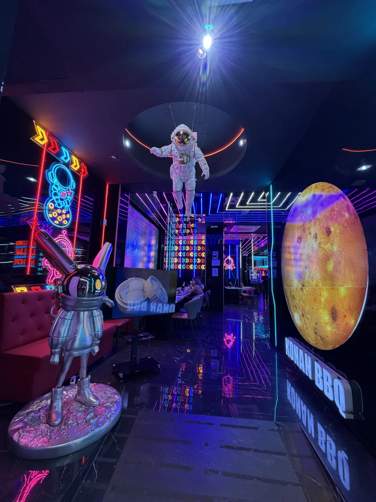 Astronaut statue suspended over the dining room at Konan BBQ, lit by arcade-style neon and a glowing planet, Málaga