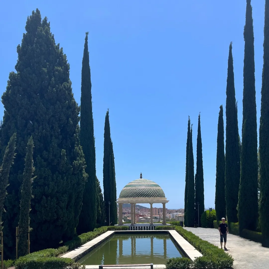 The green and white tiled pavilion at La Concepción between two rows of tall cypress trees, the long pond in front of it, Málaga's rooftops below and a man walking the gravel path on the right
