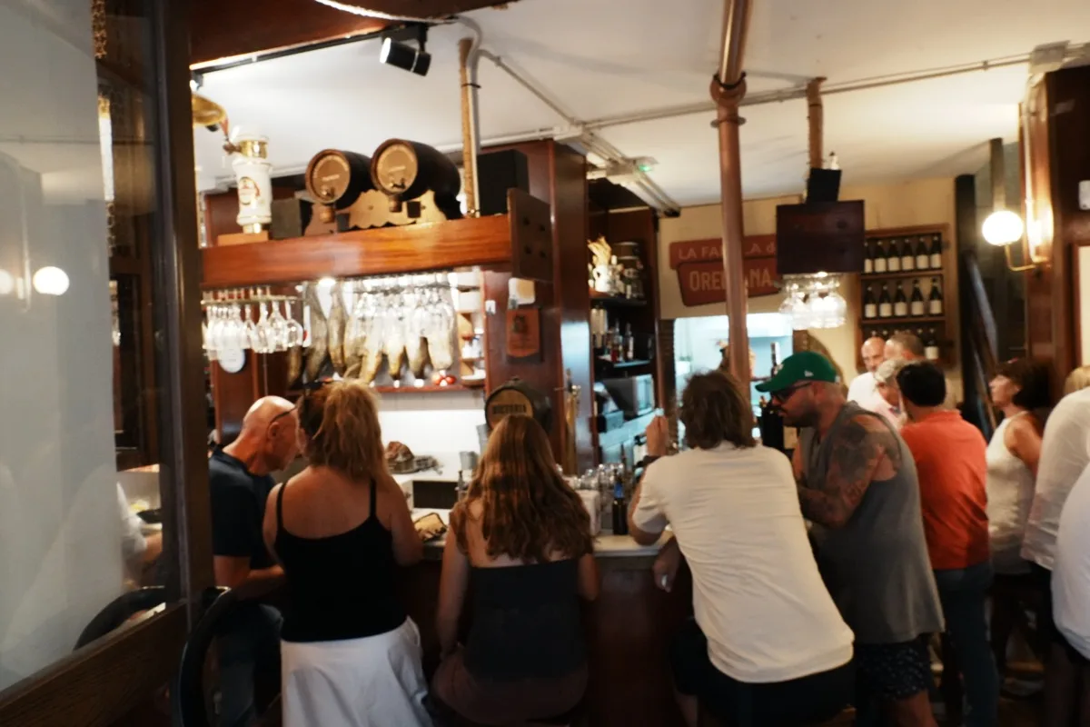 Customers standing three deep at the counter of La Farola de Orellana, hams hanging above the bar and the staff working behind it