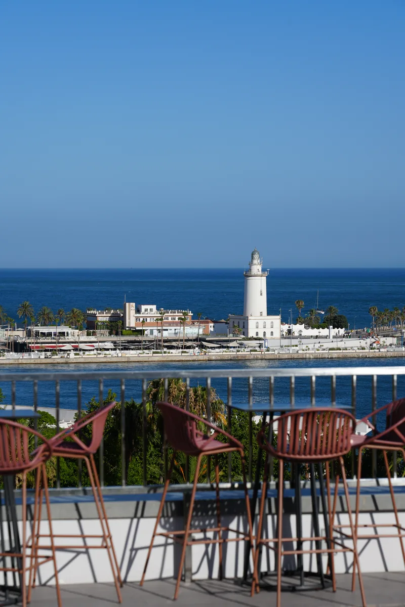 La Farola lighthouse and the port of Málaga seen from a rooftop bar terrace, red bar stools in the foreground