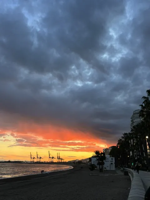 Dark storm clouds over Malagueta Beach with a band of fiery red sunset light behind the port cranes