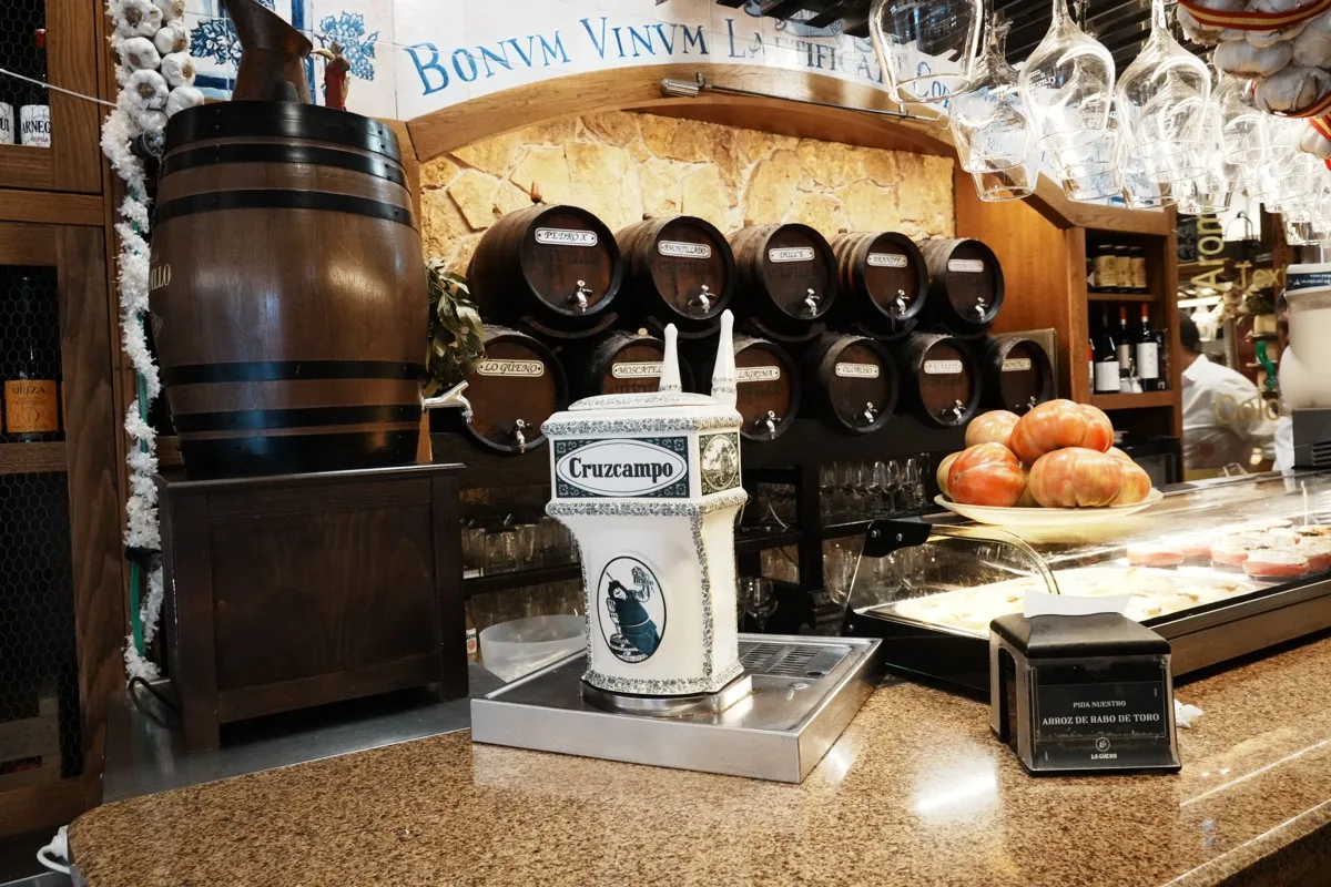 The bar at Mesón Lo Güeno, a ceramic Cruzcampo tap on the counter, wine barrels set into a stone arch under a Latin motto, garlic strings and tomatoes along the top
