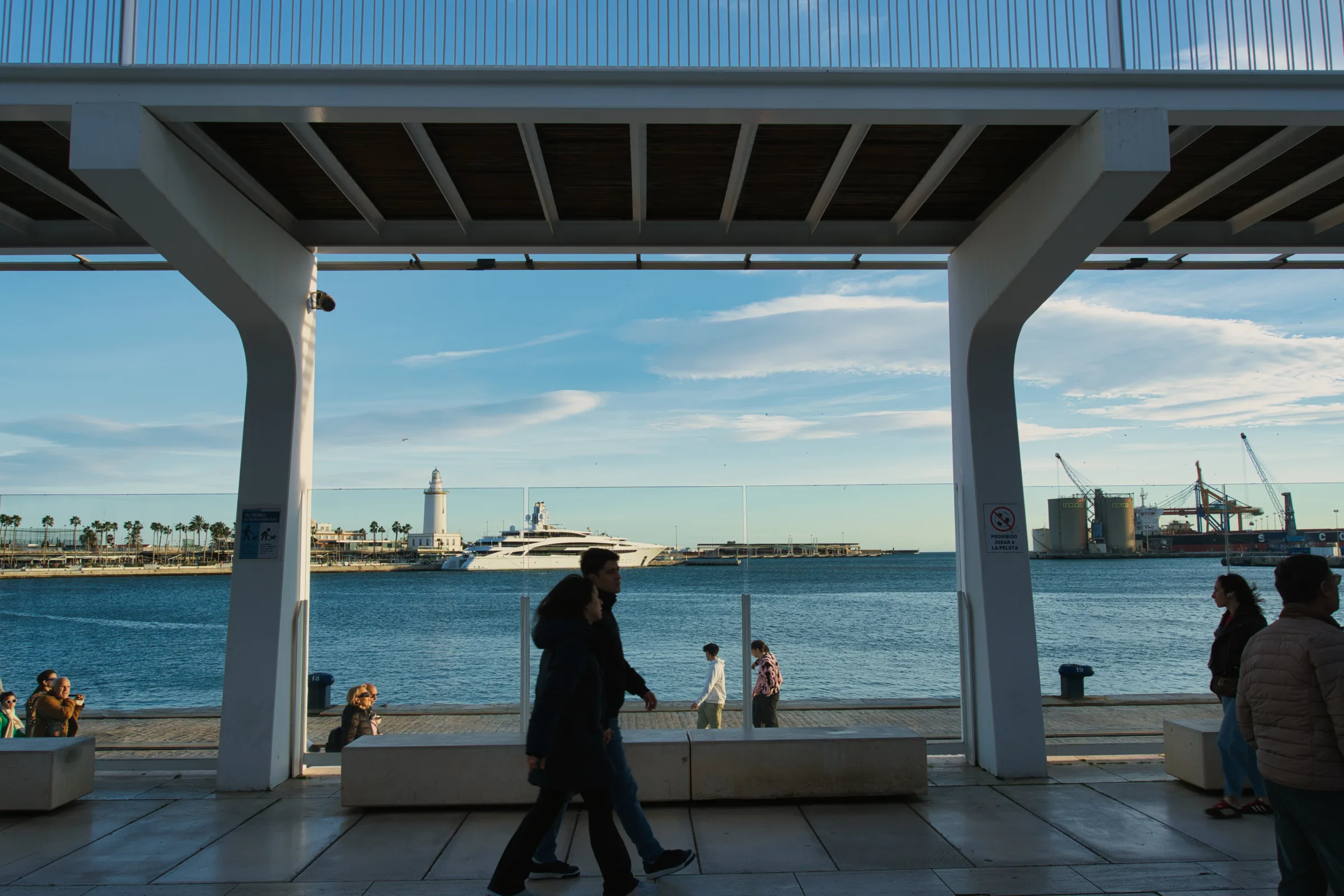 View of Malaga port from beneath Muelle Uno's wave-shaped canopy, with the harbour lighthouse in the distance